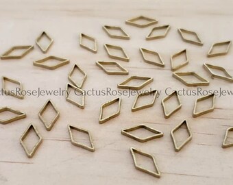 May include: A collection of small, open-frame diamond-shaped metal charms in a warm gold tone. The charms are scattered across a light-colored wooden surface, creating a delicate and artistic arrangement. These charms are ideal for jewelry making and crafting.