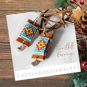 May include: A pair of handmade earrings with a Native American design. The earrings feature a rectangular leather base wrapped with silver wire and adorned with turquoise, red, and yellow beads. The earrings have silver hooks and decorative silver charms.