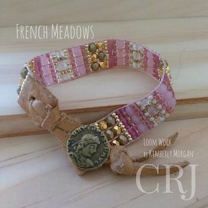 May include: A pink and gold beaded bracelet with a leather strap and a coin-shaped clasp. The bracelet is labeled "French Meadows" and "Loom Work by Kimberly Morgan CRJ".