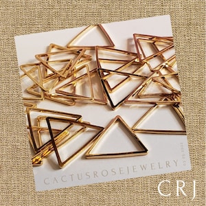 May include: A pile of gold-colored metal triangle shapes. The triangles are open on one side and have a smooth, shiny finish. The triangles are arranged in a random pattern. The text "Cactusrosejewelry" and "CRJ 03 25 2022" is visible in the bottom right corner of the image.