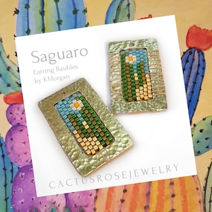 Puede incluir: Un par de pendientes de cuentas verdes, azules, amarillas y blancas con un engaste de metal martillado. Los pendientes están etiquetados como "Saguaro Earring Baubles by KMorgan" y "Cactusrosejewelry".