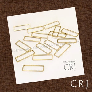 May include: A pile of 14 gold-toned metal rectangular jump rings. The rings are all the same size and shape, and they are arranged in a random pattern. The rings are on a white surface.