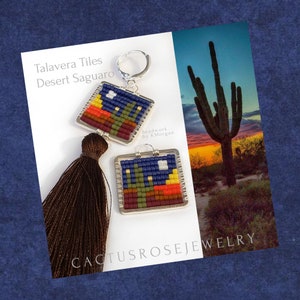 May include: A pair of beaded earrings with a southwestern desert theme. The earrings feature a square design with a blue sky, yellow moon, and a saguaro cactus in a red, orange, and green landscape. The earrings are labeled "Talavera Tiles Desert Saguaro" and "Beadwork by K.Morgan".
