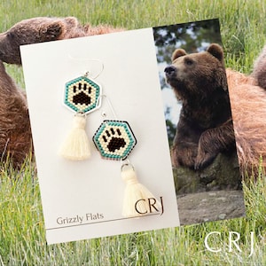 Puede incluir: Pendientes Grizzly Flats con un diseño de huella de pata de oso marrón. Los pendientes son de plata con detalles turquesa y tienen un borla blanca. El texto "Grizzly Flats" y "CRJ" es visible en la imagen.