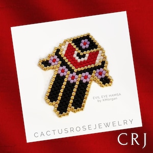 May include: A black and gold beaded hamsa hand with a red, white, and black evil eye in the center. The hamsa is surrounded by a white background with the text "EVIL EYE HAMSA by K.Morgan" and "CACTUSROSEJEWELRY CRJ".