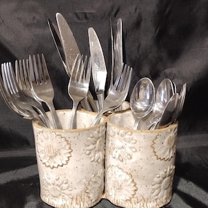 May include: A ceramic utensil holder with a cream-colored base and embossed sunflower design. The holder is divided into two sections and filled with stainless steel forks, knives, and spoons. The background is a dark fabric.