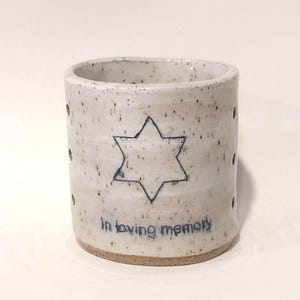 May include: A cylindrical ceramic candle holder with a speckled off-white glaze. The front features a hand-drawn six-pointed star and the text "In loving memory" in dark blue ink. Small circular holes are spaced vertically along the sides.