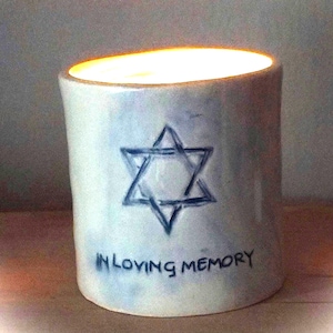 May include: A cylindrical ceramic candle holder with a glossy white finish and a blue-toned wash. The front features a hand-drawn Star of David in blue ink above the text IN LOVING MEMORY. The interior glows with a warm, bright light.