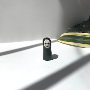 May include: A black and white clay figurine of No Face from the animated film Spirited Away. The figurine is standing on a white surface.