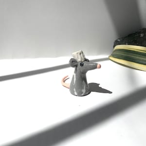 May include: A small grey ceramic mouse figurine wearing a white ceramic hat. The mouse has a pink nose and black eyes.