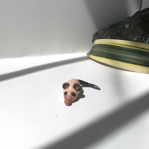 May include: A small, white ceramic mouse figurine with black ears and a pink nose. The mouse is lying on its belly with its tail curled up.
