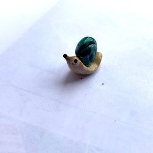 May include: A small, hand-crafted ceramic snail figurine. The snail is painted in shades of green and beige.