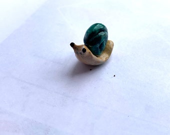 May include: A small, hand-crafted ceramic snail figurine. The snail is painted in shades of green and beige.