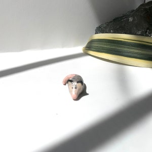 May include: A small ceramic figurine of a gray and white opossum with a pink nose and tail.