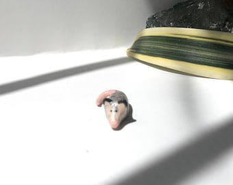 May include: A small ceramic figurine of a gray and white opossum with a pink nose and tail.