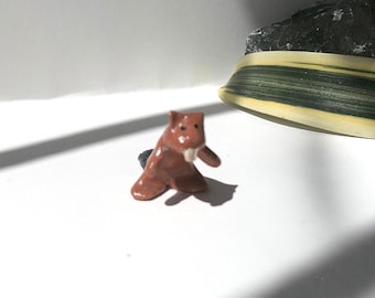 May include: A small, brown ceramic figurine of a beaver with white teeth. The beaver is standing on a white surface.