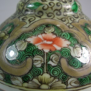 Pode incluir: Close-up de um objeto cer&acirc;mico decorativo com um design floral. O objeto apresenta uma flor laranja central rodeada por espirais e folhas verdes, sobre um fundo creme e dourado. O design &eacute; detalhado e ornamentado.