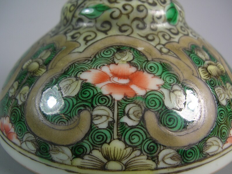 Pode incluir: Close-up de um objeto cer&acirc;mico decorativo com um design floral. O objeto apresenta uma flor laranja central rodeada por espirais e folhas verdes, sobre um fundo creme e dourado. O design &eacute; detalhado e ornamentado.