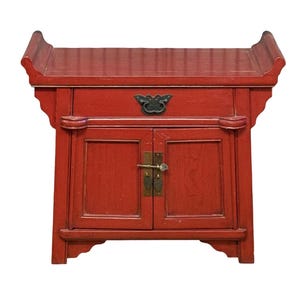 May include: A red wooden cabinet featuring a flat top with curved, flared ends. The cabinet includes one upper drawer with a dark metal butterfly-shaped pull and two lower doors secured by a brass latch mechanism with a small key attached.