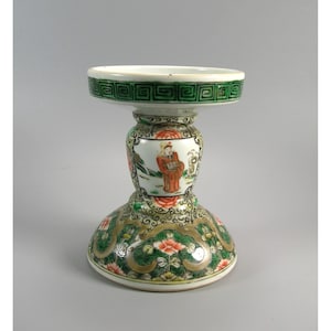 Pode incluir: Um pedestal decorativo de porcelana em forma de ampulheta. A base e o topo s&atilde;o brancos com padr&otilde;es florais e geom&eacute;tricos verdes e dourados. O centro apresenta uma cena pintada de uma pessoa em trajes tradicionais.