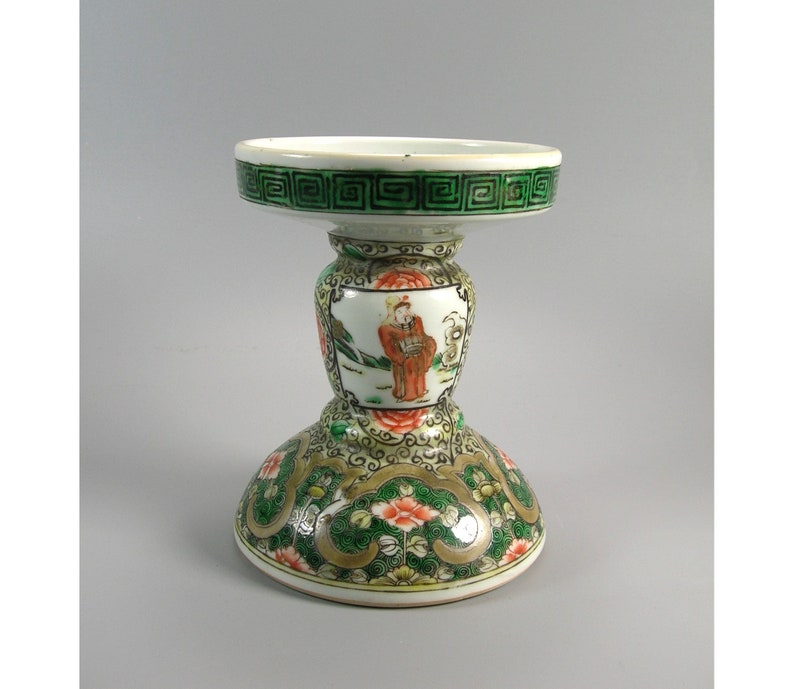 Pode incluir: Um pedestal decorativo de porcelana em forma de ampulheta. A base e o topo s&atilde;o brancos com padr&otilde;es florais e geom&eacute;tricos verdes e dourados. O centro apresenta uma cena pintada de uma pessoa em trajes tradicionais.
