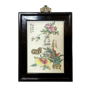 May include: A framed, hand-painted artwork featuring a white vase with pink flowers, peaches, and coins. The artwork is set in a black wooden frame with a decorative brass accent at the top. The painting has Chinese characters.