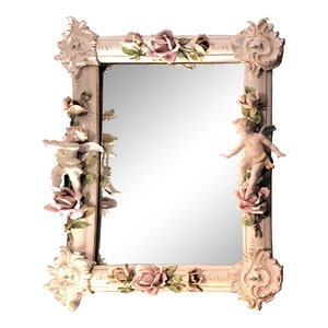 Könnte beinhalten: Ein weißer Porzellan-Spiegelrahmen mit je einem Cherub auf jeder Seite, umgeben von rosa Rosen und grünen Blättern.