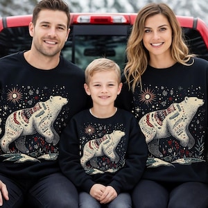 May include: Black sweatshirts featuring a bear design. A family of three and a golden retriever dog are sitting in the back of a red truck. The bear design is white with colourful patterns. The word "Black" is printed on the truck.
