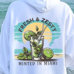 Turn up the tropical energy with our Fresh & Zesty – Minted in Miami hoodie, featuring a bold mojito graphic set against a dreamy Miami sunset beach scene. This eye-catching back print channels sunny coastal vibes, citrusy freshness, and laid-back luxury—perfect for sipping mojitos or dreaming of South Beach. A go-to for travel, lounging, or gifting anyone who loves cocktails, sunshine, and Miami heat.  Ideal gift for cocktail lovers, beachgoers, or anyone with a thing for Miami vibes.