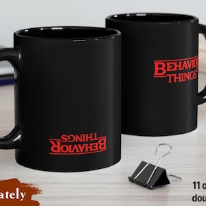 May include: Two black ceramic mugs with a glossy finish, each featuring the red text "BEHAVIOR THINGS" printed in a stylized font. The mugs have a standard handle and a cylindrical shape, with the text appearing on both sides of the ceramic surface.