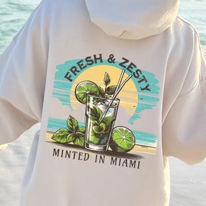 Turn up the tropical energy with our Fresh & Zesty – Minted in Miami hoodie, featuring a bold mojito graphic set against a dreamy Miami sunset beach scene. This eye-catching back print channels sunny coastal vibes, citrusy freshness, and laid-back luxury—perfect for sipping mojitos or dreaming of South Beach. A go-to for travel, lounging, or gifting anyone who loves cocktails, sunshine, and Miami heat.  Ideal gift for cocktail lovers, beachgoers, or anyone with a thing for Miami vibes.