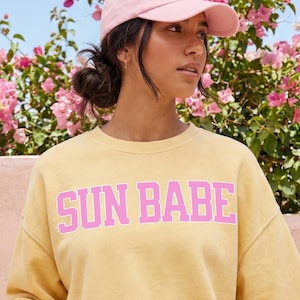 May include: A pale yellow crewneck sweatshirt featuring the text SUN BABE in large pink block letters across the chest. The garment is worn with a light pink baseball cap and a pink tumbler with a straw is held in one hand. The design is printed.