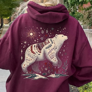 May include: Maroon coloured hoodie with a detailed illustration of a bear. The bear is white with red and cream patterns, set against a backdrop of stars and a sun. The word "Maroon" is printed at the bottom of the image.