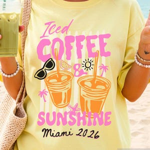 May include: A pale yellow t-shirt with the text "Iced Coffee & Sunshine Miami 2026" in pink and black. The design features sunglasses, coffee cups, and a sun. The person is holding a green drink in a clear cup with a straw.