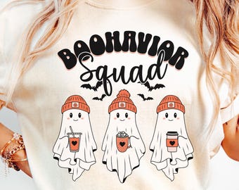 May include: Ivory-colored t-shirt with the text "BOOHAVIOR Squad" above three cartoon ghosts wearing orange beanies. Each ghost holds a beverage: a drink with a straw, a mug with hearts, and a coffee cup.