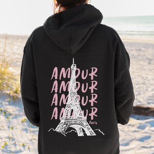 K&ouml;nnte beinhalten: Ein schwarzer Kapuzenpullover mit einer wei&szlig;en Grafik des Eiffelturms auf der R&uuml;ckseite. Das Wort AMOUR ist viermal vertikal in Hellrosa hinter dem Turm aufgedruckt, wobei das Wort Paris in kleiner wei&szlig;er Schrift nahe der Basis steht.