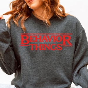 May include: A dark heather gray crewneck sweatshirt featuring the red text "BEHAVIOR THINGS" printed on the chest. The text is stylized with horizontal lines above and below the word "BEHAVIOR" and is centered on the front of the garment.