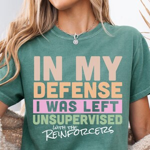 May include: Light green t-shirt featuring the text "IN MY DEFENSE I WAS LEFT UNSUPERVISED WITH THE REINFORCERS" in multiple colours. The shirt has a crew neck and short sleeves.