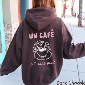 May include: A dark chocolate brown hooded sweatshirt featuring a graphic on the back. The design includes a white line drawing of a croissant resting on a coffee cup and saucer, with the text UN CAF&Eacute; and s'il vous pla&icirc;t in light pink.