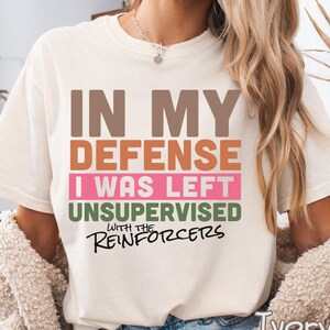 May include: Ivory-colored t-shirt with the text "IN MY DEFENSE I WAS LEFT UNSUPERVISED WITH THE REINFORCERS" in brown, orange, pink, and green. The shirt is a relaxed fit.