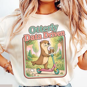 May include: An ivory t-shirt featuring a graphic of a brown otter riding a pink and blue scooter while holding a coffee cup. The text above the illustration reads "Otterly Data Driven" in green and pink letters, set against a forest background.