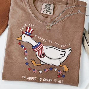 May include: Brown t-shirt with a graphic of a goose wearing a patriotic hat and scarf, with the text "FROM THE WINDOWS TO THE WALLS" and "I'M ABOUT TO GRAPH IT ALL". The shirt has a crew neck and short sleeves.