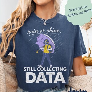 May include: Denim blue t-shirt with the text "rain or shine" and "Still Collecting Data." The graphic shows a person under a purple umbrella. The shirt also has a small text bubble that says "Great gift for BCBA's and RBT'S."