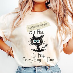 May include: Ivory-colored t-shirt with the text "Extinction Burst in Progress" and "It's Fine, I'm Fine, Everything Is Fine." A distressed black cat graphic with wide blue eyes is featured below the text. The shirt is made of a soft material.