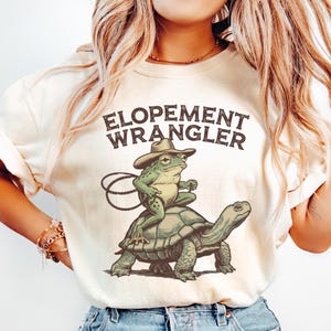 May include: Ivory-colored t-shirt featuring the text "ELOPEMENT WRANGLER" above an illustration of a frog wearing a cowboy hat riding a turtle. The frog holds a rope, and the shirt is made of a soft material.