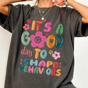 May include: Dark grey t-shirt featuring the phrase "IT'S A GOOD day TO SHAPE BEHAVIORS" in vibrant, multi-coloured letters. The design incorporates floral and heart motifs. A relaxed, comfortable style of top.