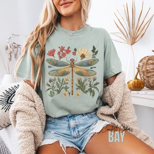 This Women's Dragonfly Floral T-Shirt is a boho and nature-inspired tee that is perfect for garden lovers and those who embrace the cottagecore aesthetic. It exudes a peaceful and whimsical vibe, making it a great addition to a nature lover's wardrobe. Ideal for casual wear or as a thoughtful gift for a loved one who enjoys botanical designs. Relevant for Earth Day, birthdays, and any nature-themed celebrations.