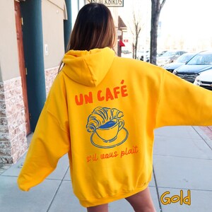 May include: A bright yellow hooded sweatshirt featuring a graphic on the back. The design includes the orange text "UN CAF&Eacute;" above a blue line drawing of a croissant resting on a coffee cup and saucer, with the orange text "s'il vous plait" below.