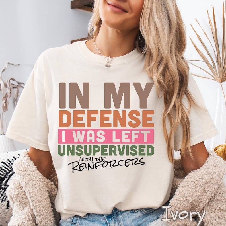 May include: Ivory-colored t-shirt with the text "IN MY DEFENSE I WAS LEFT UNSUPERVISED WITH THE REINFORCERS" in brown, orange, pink, and green. The shirt is a relaxed fit.