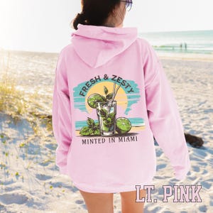 Turn up the tropical energy with our Fresh & Zesty – Minted in Miami hoodie, featuring a bold mojito graphic set against a dreamy Miami sunset beach scene. This eye-catching back print channels sunny coastal vibes, citrusy freshness, and laid-back luxury—perfect for sipping mojitos or dreaming of South Beach. A go-to for travel, lounging, or gifting anyone who loves cocktails, sunshine, and Miami heat.  Ideal gift for cocktail lovers, beachgoers, or anyone with a thing for Miami vibes.